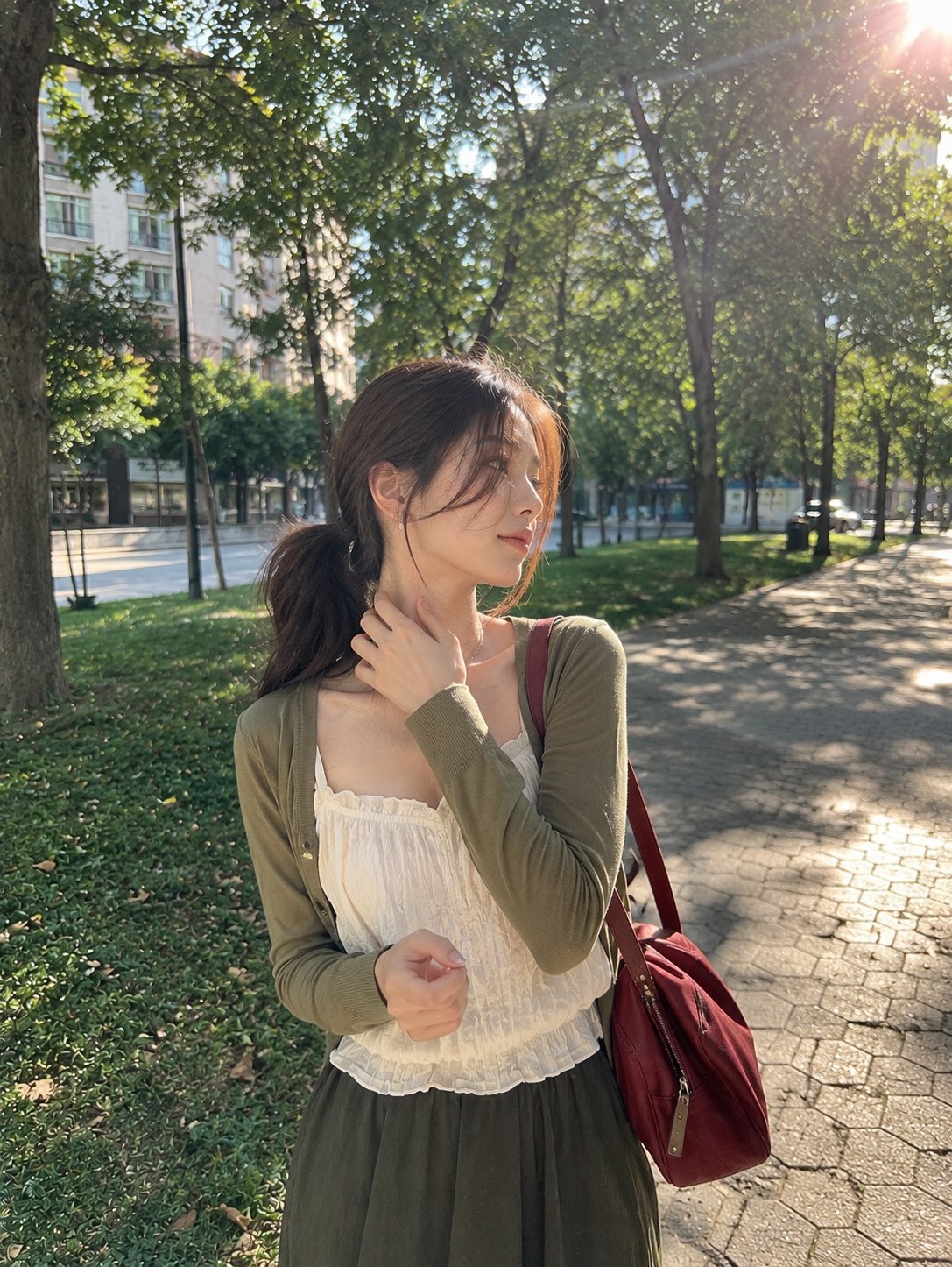 一张写实风格的户外抓拍人像照片，一位年轻女性站在午后阳光下的宁静城市公园中。她留着深棕色长发，松松地扎成低马尾，几缕轻盈的发丝自然垂落在脸庞两侧。她目光柔和地望向一侧，神情平静而若有所思。

她身穿一 — AI image prompt — GPT Image 2